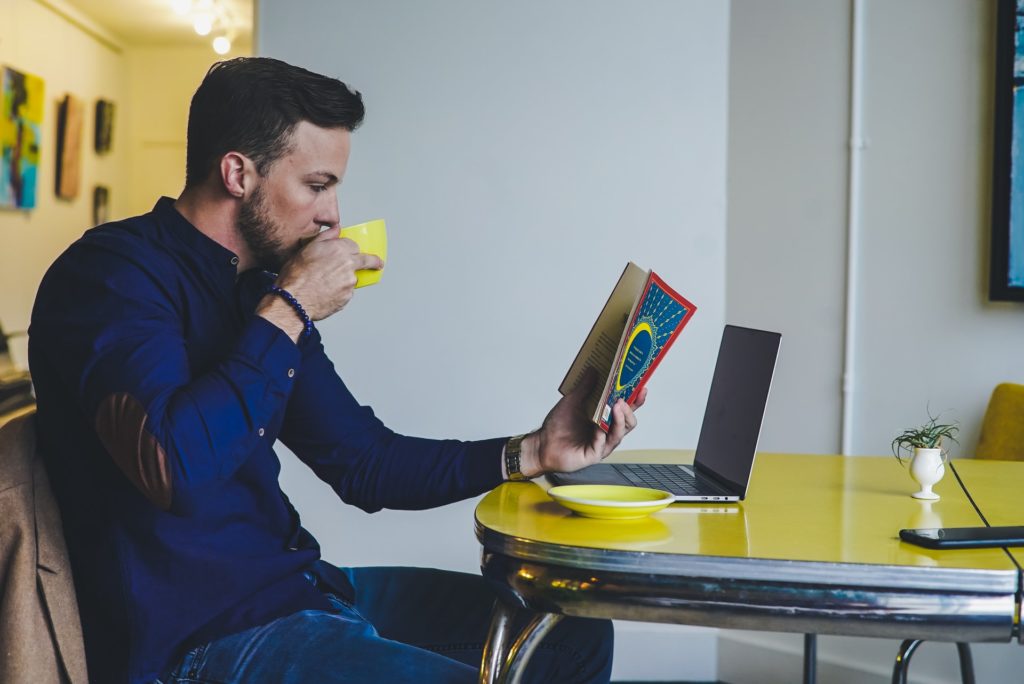 Homme lisant livre et buvant dans une tasse devant un ordinateur